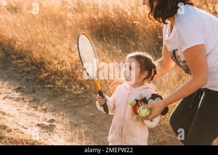 Das süße kleine Mädchen genießt einen Tennisschläger in der Hand. Mom gibt ihr einen Tennisball Stockfoto