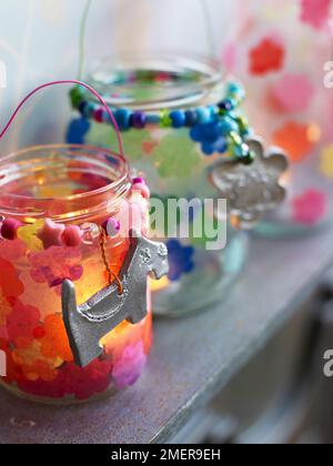 Gläser aus Kerzenglas mit Papiertaschenblumen und silbernen Anhängern auf dem Kaminsims Stockfoto