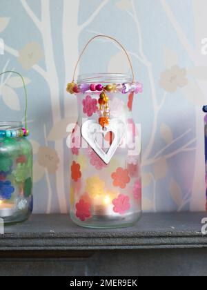 Gläser aus Kerzenglas mit Papiertaschenblumen und silbernen Anhängern auf dem Kaminsims Stockfoto