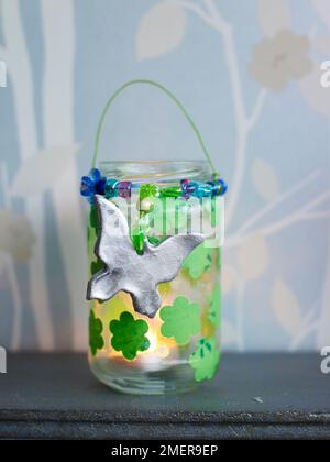 Gläser aus Kerzenglas mit Papiertaschenblumen und silbernen Anhängern auf dem Kaminsims Stockfoto