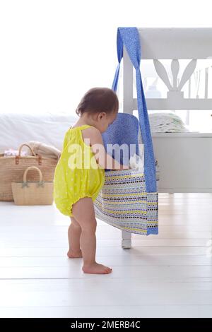 Ein kleines Mädchen, das in eine Windeltasche schaut, die an der Rückseite des Stuhls hängt, 13 Monate Stockfoto