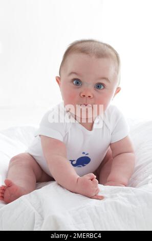 Kleiner Junge, 7 Monate, sitzt auf einem Bett, trägt einen schablonierten Strampler Stockfoto