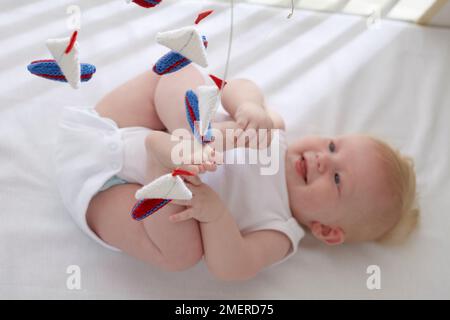 Ein Junge, der sich hinlegt und bis zum gestrickten Segelboot-Mobile greift, 5 Monate Stockfoto