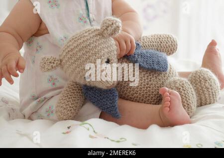 Kleines Mädchen, das mit gestricktem Teddybär auf dem Schoß spielt, 9 Monate Stockfoto