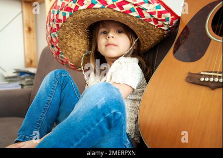 Süßes kleines Mädchen in lässiger Kleidung und farbenfroher Sombrero, der zu Hause auf dem Sofa neben der Gitarre sitzt. Kaukasische kleine Mädchen trägt mexikanischen Stil Stroh Stockfoto