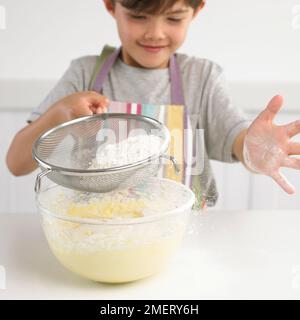 Ein Junge siebt Mehl über eine Schüssel Kuchenmischung, 7 Jahre Stockfoto