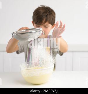 Ein Junge siebt Mehl über eine Schüssel Kuchenmischung, 7 Jahre Stockfoto