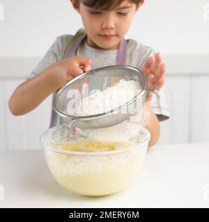 Ein Junge siebt Mehl über eine Schüssel Kuchenmischung, 7 Jahre Stockfoto