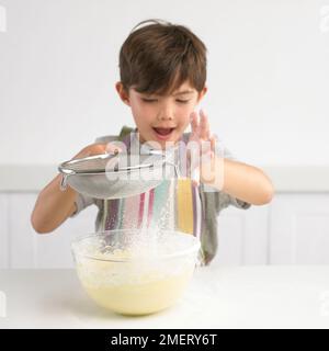 Ein Junge siebt Mehl über eine Schüssel Kuchenmischung, 7 Jahre Stockfoto