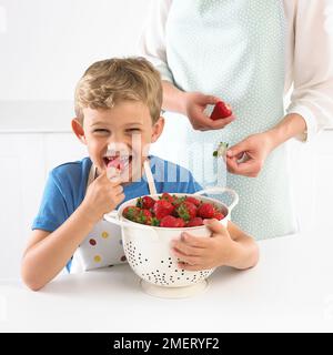 Ein Junge, der ein Sieb voll hält und eine Erdbeere isst, 6 Jahre Stockfoto