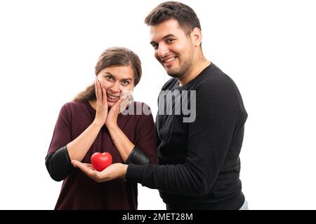 Ein lächelnder Freund, der seiner süßen Freundin ein rotes Herz schenkt, mit Liebe, die einen überraschenden Wow-Ausdruck und eine Geste auf weißem Studiohintergrund macht Stockfoto