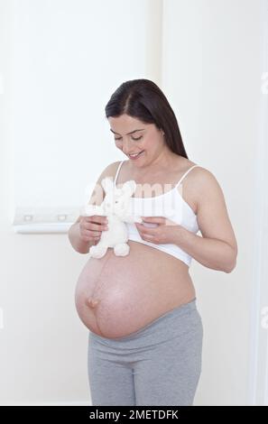 Schwer schwangere Frau, deren Bauch freigelegt ist und ein weiches Spielzeug hält Stockfoto