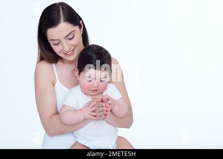 Kleines Mädchen (40 Wochen), das auf dem Schoß seiner Mutter sitzt und die Hände ihrer Mutter berührt Stockfoto