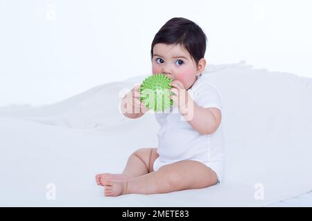 Ein kleines Mädchen (40 Wochen) hält einen Gummiball an den Mund Stockfoto