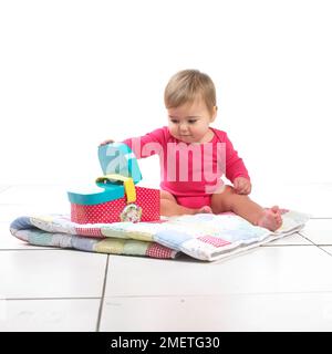 Kleines Mädchen (12 Monate), das auf einer Steppdecke sitzt und mit einem Spielzeug-Picknickkorb spielt Stockfoto