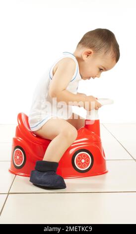 Ein Junge mit Weste und Pantoffeln sitzt auf einem roten Autotopf mit Lenkrad, 15 Monate Stockfoto