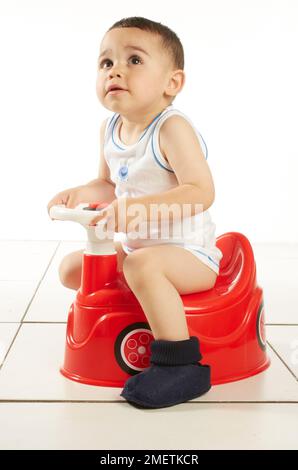 Ein Junge mit Weste und Pantoffeln sitzt auf einem roten Autotopf mit Lenkrad, 15 Monate Stockfoto