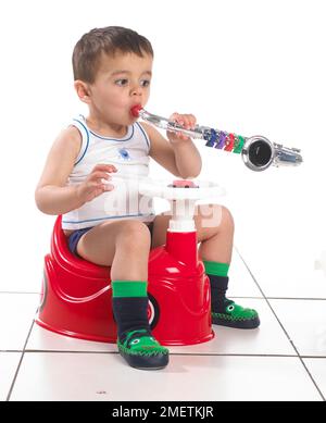 Ein Junge mit Weste und Hausschuhen sitzt auf einem Töpfchen in der Form eines Autos mit Lenkrad und hält ein Saxophon, 15 Monate Stockfoto