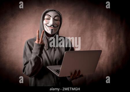 Hacker mit anonymer Maske mit einem Laptop, der das Siegessymbol macht, Studiofoto Stockfoto