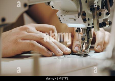 Maßgeschneidertes Nähen am Arbeitsplatz. Männerhände nähen in seinem Studio an der Maschine an. Maßgeschneidertes Konzept. Nahaufnahme von Nadel und Faden in männlichen Händen Stockfoto