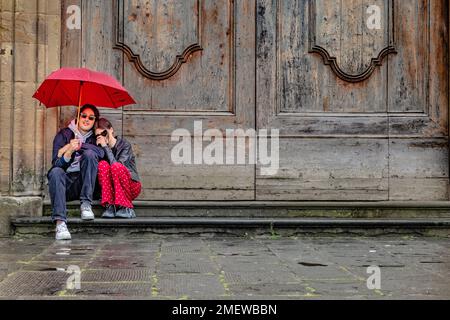 Ein junges Paar, das vor der Tür unter einem roten Schirm sitzt, in Florenz, Italien. Stockfoto