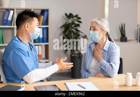 Seriöse reife koreanische Ärztin in Schutzmaske konsultiert alte europäische Patientin in der Klinik Stockfoto