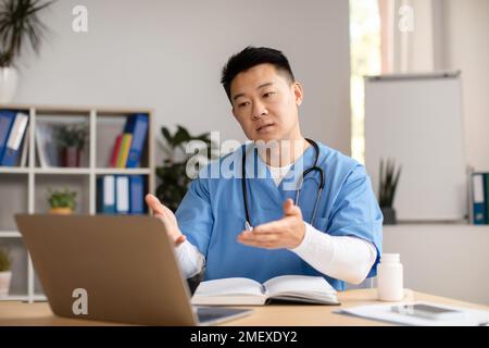 Seriöser, reifer chinesischer Therapeut in Uniform, Computer anschauen, sprechen, Gesten am Tisch in der Klinik Stockfoto