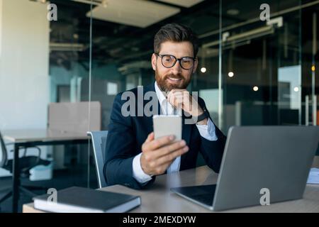 Geschäftskonzept. Positiver Unternehmer mittleren Alters, der ein Smartphone und einen modernen Laptop benutzt, sitzt am Schreibtisch im Büro Stockfoto