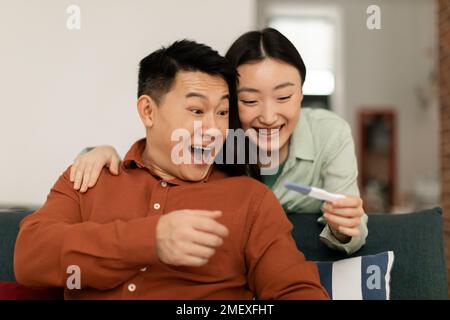 Überfreuliche asiatische Frau, die positive Ergebnisse des Schwangerschaftstests zeigte, an überraschten Ehemann, sitzend auf der Couch, Kopierraum Stockfoto