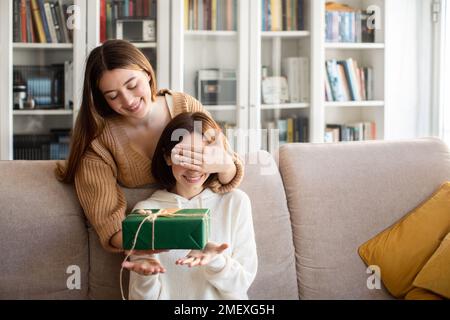 Die fröhliche europäische Millennials-Dame schließt die Augen vor ihrem Freund und schenkt ihr eine Geschenkbox im Wohnzimmer Stockfoto