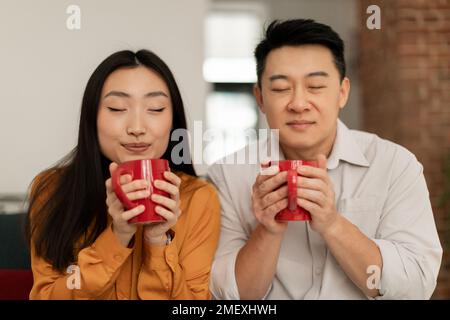 Junge asiatische Frau und reifer Mann riechen frischen aromatischen Kaffee mit geschlossenen Augen und genießen ein heißes Morgengetränk Stockfoto