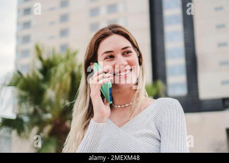 Junge, hübsche junge Frau, die mit einem Smartphone lächelt und spricht. Glückliche blonde Dame, die einen Anruf in der City City City Street hat. Kommunikationskonzept. Hochwertiges Foto Stockfoto