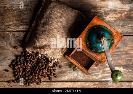 Draufsicht auf eine antike Kaffeemühle mit offener Schublade, die gemahlenen Kaffee, Esparto-Sack und verstreute Kaffeebohnen auf einem rustikalen Holztisch zeigt. Stockfoto