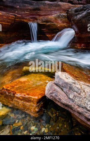 Blaue Wasserfälle im Glacier-Nationalpark vor den roten und orangenen Felsen an der Going to Sun Road in Montana. Stockfoto