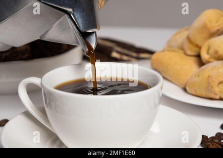 Der Vorgang, bei dem Kaffee aus einer Geysir-Moka-Kaffeemaschine in eine weiße Tasse gegossen wird Stockfoto