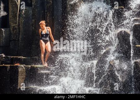 Weiße Frau in schwarzem Badeanzug erfrischt sich unter fallenden Wasserströmen, die über schwarze Vulkansteinkaskaden fließen. Wasserfall Rochester Falls - Popul Stockfoto