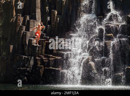 Ein Mann mittleren Alters, der in einen Wasserfallsee springt. Fallende Wasserströme fließen auf schwarzen Vulkansteinkaskaden. Wasserfall Rochester Falls - beliebter Touristenbereich Stockfoto