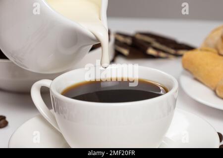 Die Sahne aus dem Milchkännchen wird in den Kaffee gegossen, wobei Kaffee mit Sahne und Cappuccino zubereitet wird. Stockfoto