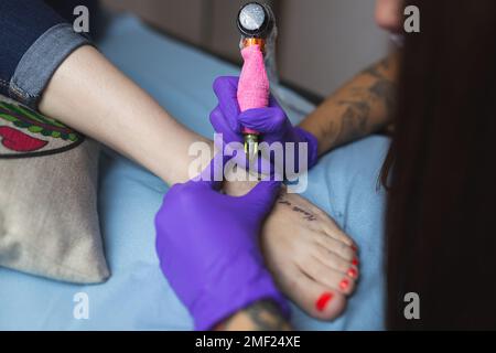 Eine Tätowierungskünstlerin, die einen Fuß tätowiert. Tätowierung eines Satzes am Fuß, pinkfarbene Tattoo-Maschine. Stockfoto