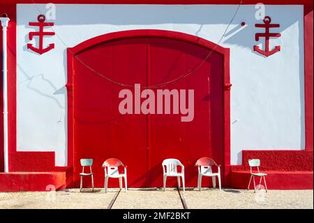 Rote Doppeltüren und weißes Gebäude mit Stühlen im Freien. Altes Gebäude der Rettungsschwimmerstation aus den 1920er Jahren in Alvor, Algarve, Portugal. Stockfoto