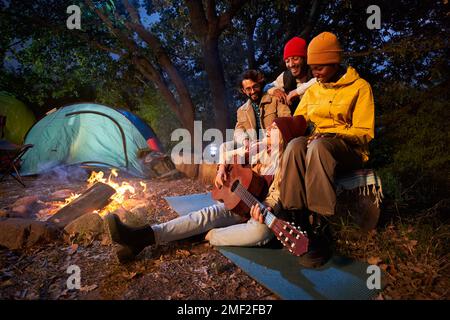 Gruppe von Freunden mit Gitarre am Lagerfeuer und Campingzelt im Freien am Nachmittag. Stockfoto