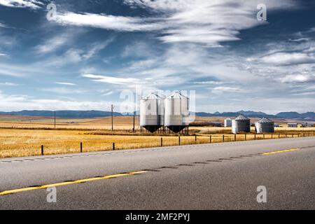 Ein Paar Silos aus Stahlkorn, die neben einer geteilten Autobahn entlang der ernteten Felder und der fernen Rocky Mountains im Hintergrund in der Nähe von Longview, Alberta, Kalifornien, stehen Stockfoto