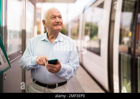 Älterer Mann mit Smartphone, während er auf der Straßenbahnhaltestelle steht Stockfoto