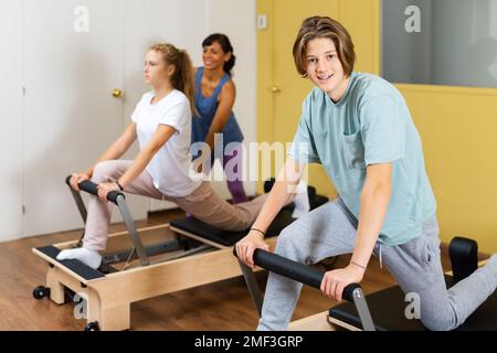 Kleiner Junge, der in der Kamera schaut, während er Pilates im Fitnessstudio trainiert. Ihre Trainerin hispanische Frau hilft Mädchen mit Übungen auf Pilates Reformer im Hintergrund. Stockfoto