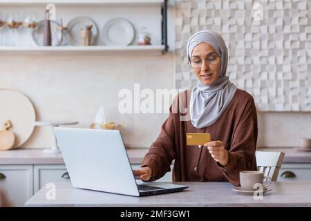 Fröhliche und erfolgreiche Frau in Hidschab zu Hause in der Küche mit Laptop und Bankkreditkarte kauft online im Online-Shop ein, muslimische Frau wählt Produkte und Geschenke aus der Ferne. Stockfoto