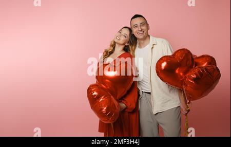 Ein Paar, rote Ballons in Form eines Herzens auf pinkfarbenem Hintergrund. Valentinstag, Liebe, 14. Februar. Eine Frau in einem roten Kleid lächelt in die Kamera Stockfoto