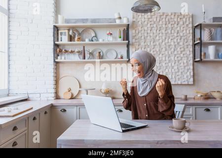 Freiberuflerin in Hijab, die von zu Hause aus an einem Laptop arbeitete, erhielt Frau gute Nachrichten und gute Nachrichten online-Benachrichtigung, glücklich und feierte gute Erfolge, Geschäftsfrau im Home Office. Stockfoto