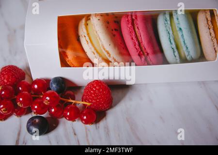 Macaron Dessert, eine Schachtel mit bunten Makronen, liegt auf einem Marmorstil. Süßigkeiten, Diät, Geschenk für den 8. März oder 14. Februar. Erdbeeren und Preiselbeeren, b Stockfoto