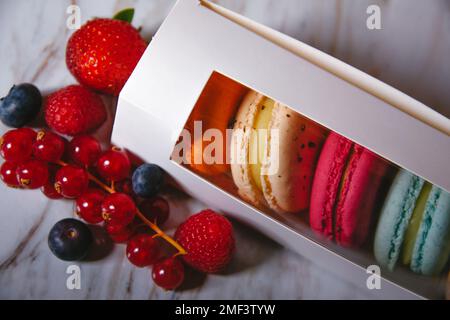 Makaronis in einem weißen Kasten mit transparentem Fenster. Farbdessert, rosafarbene, mintfarbene, blaue und orangefarbene Makronen. Erdbeeren, Himbeeren, Heidelbeeren und Stockfoto