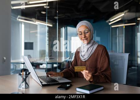 Eine junge muslimische Frau in einem Hidschab sitzt im Büro an einem Tisch mit einem Laptop, hält eine Kreditkarte und benutzt sie. Bezahlt Rechnungen, Schecks, Online-Shopping. Stockfoto